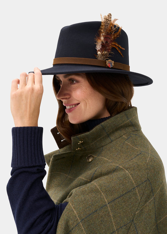 Navy felt unisex hat with brown leather band and feather embellishment by Richmond, worn in side view with olive plaid coat and navy sweater.