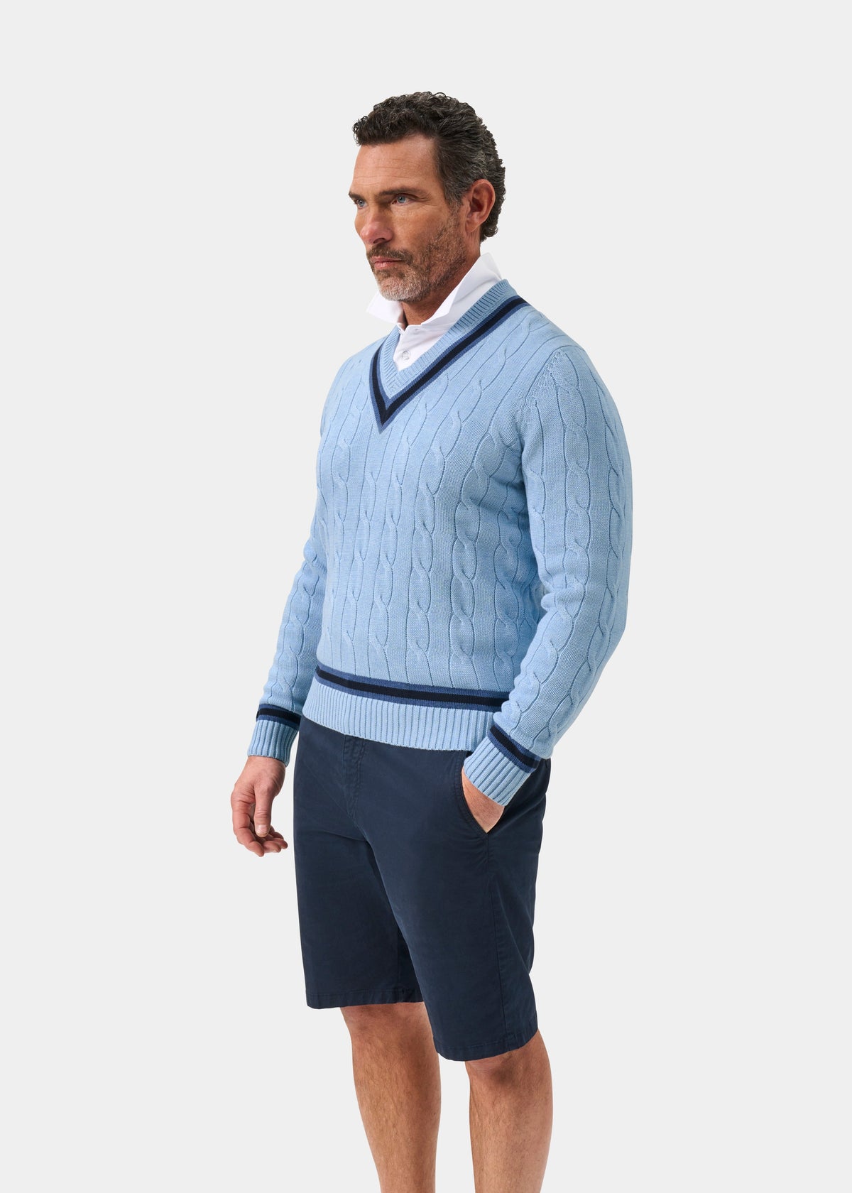 Steel blue cotton cable knit V-neck jumper with dark navy trim by Sandridge, shown on model in side view with white shirt and navy shorts