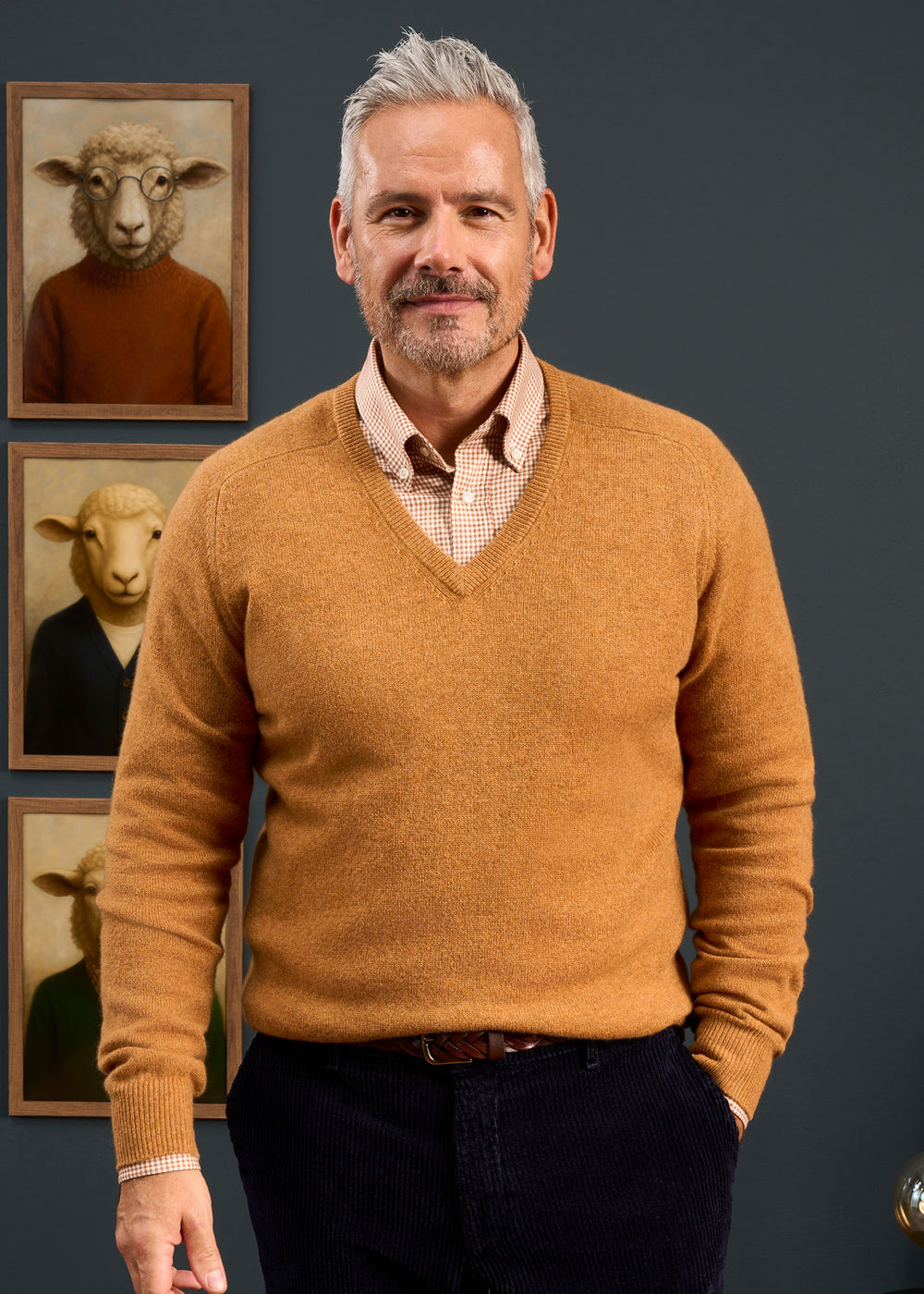 Smiling man in a Hampshire Lambswool Vee Neck Jumper (Gazelle) stands before framed sheep portraits on dark wall.