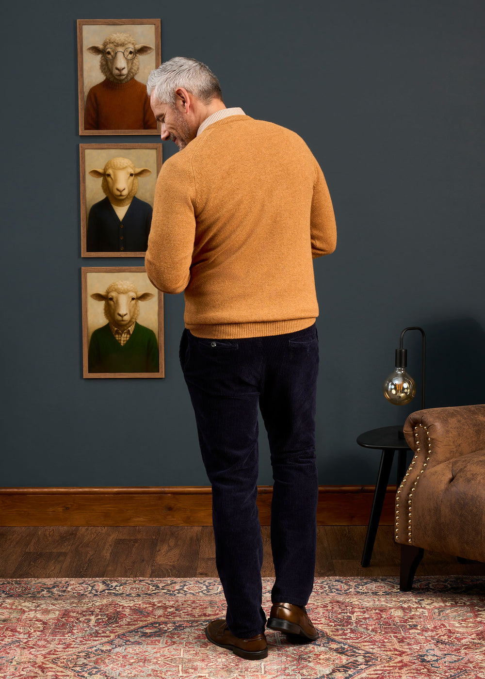 Man wears Hampshire Lambswool Vee Neck Jumper in Gazelle, standing before three sheep portraits on a dark wall.