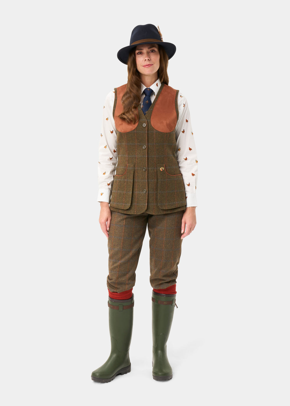 Woman models Combrook Ladies Tweed Shooting Waistcoat in Willow with shirt, tie, hat, green boots, and brown pants.