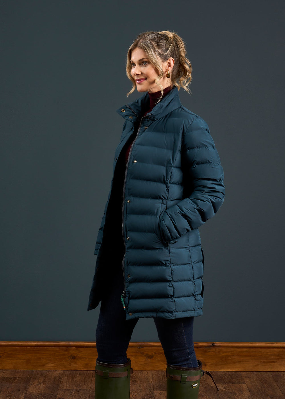 Woman in a Calsall Ladies Coat in Navy - Regular Fit, dark pants, and green boots stands sideways on a dark background.
