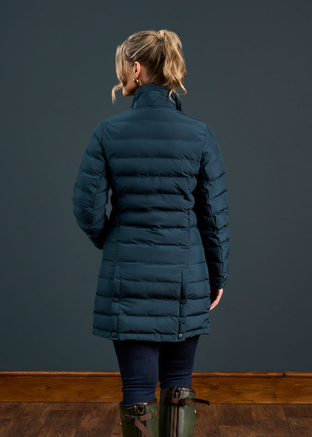 Woman with blonde ponytail in a Calsall Ladies Coat in Navy - Regular Fit, jeans, and boots, facing a dark wall.