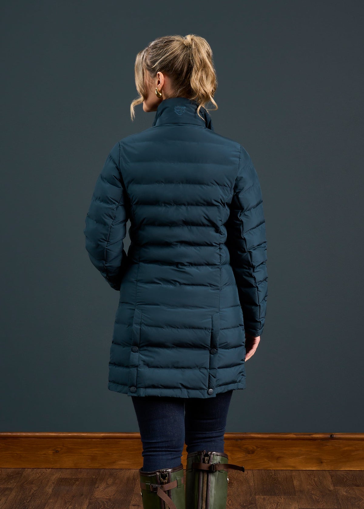 Woman with blonde ponytail in a Calsall Ladies Coat in Navy - Regular Fit, jeans, and boots, facing a dark wall.