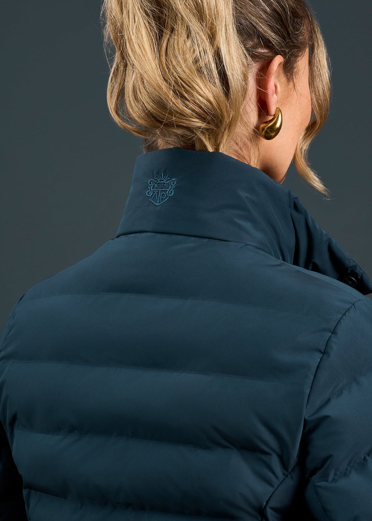 Blonde woman in ponytail, facing away, wears Calsall Ladies Coat in Navy - Regular Fit and gold hoop earrings.