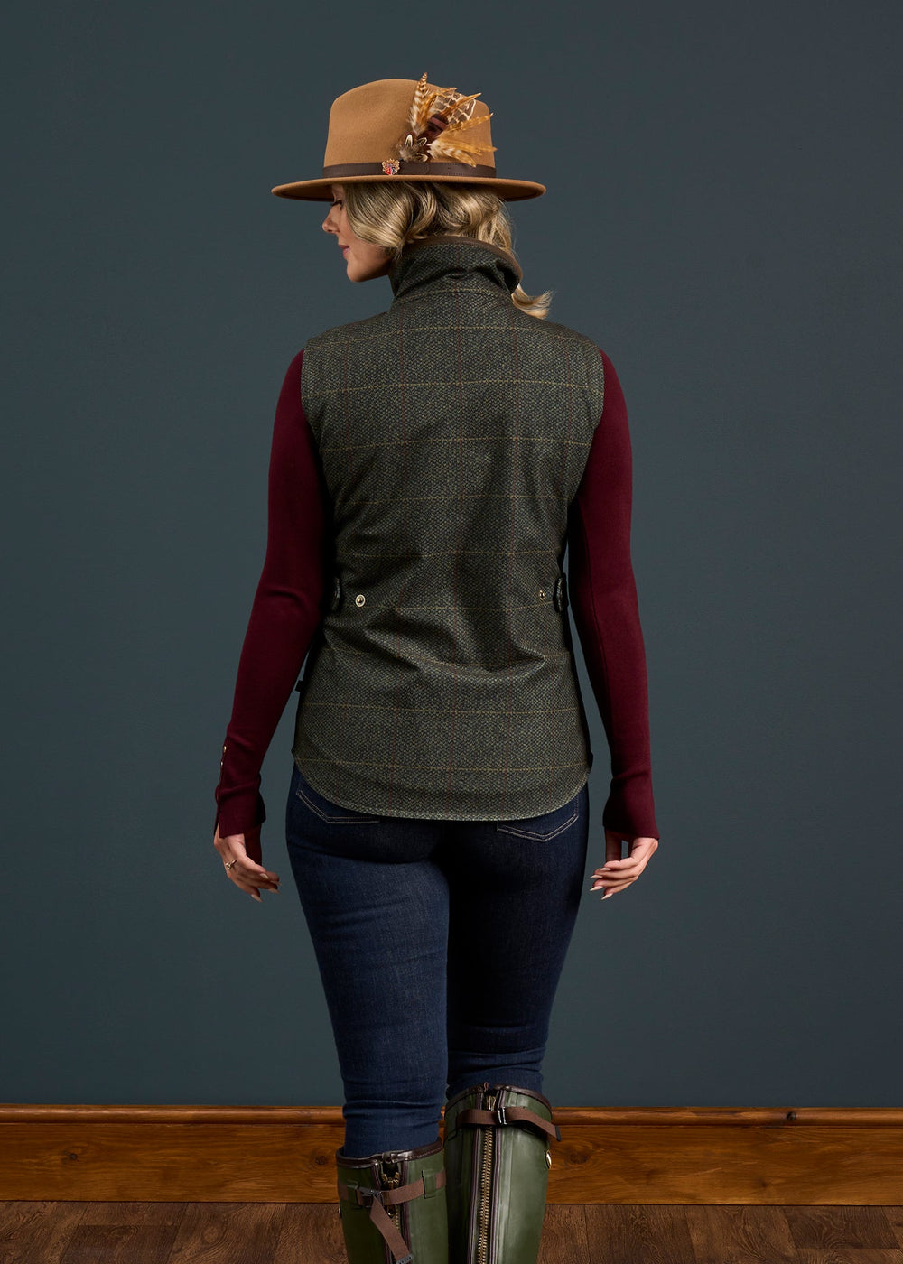 Woman facing away, wearing brown feathered hat, maroon sleeves, jeans, green boots & Didsmere Myrtle Gilet.