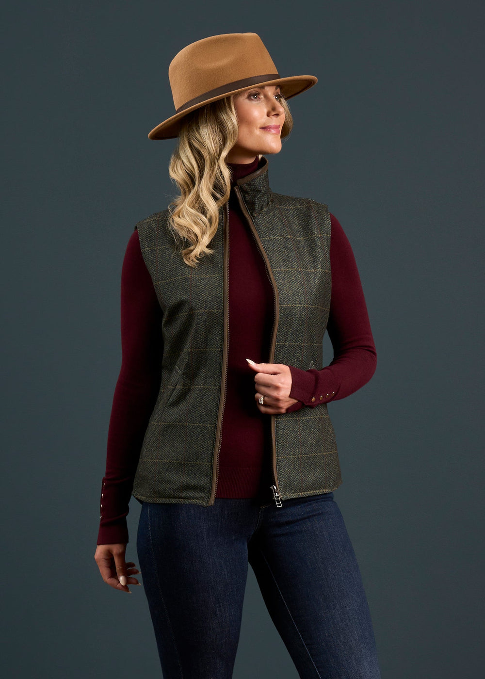 Woman in brown hat, Didsmere Ladies gilet in Myrtle, burgundy turtleneck, and jeans poses on dark gray backdrop.