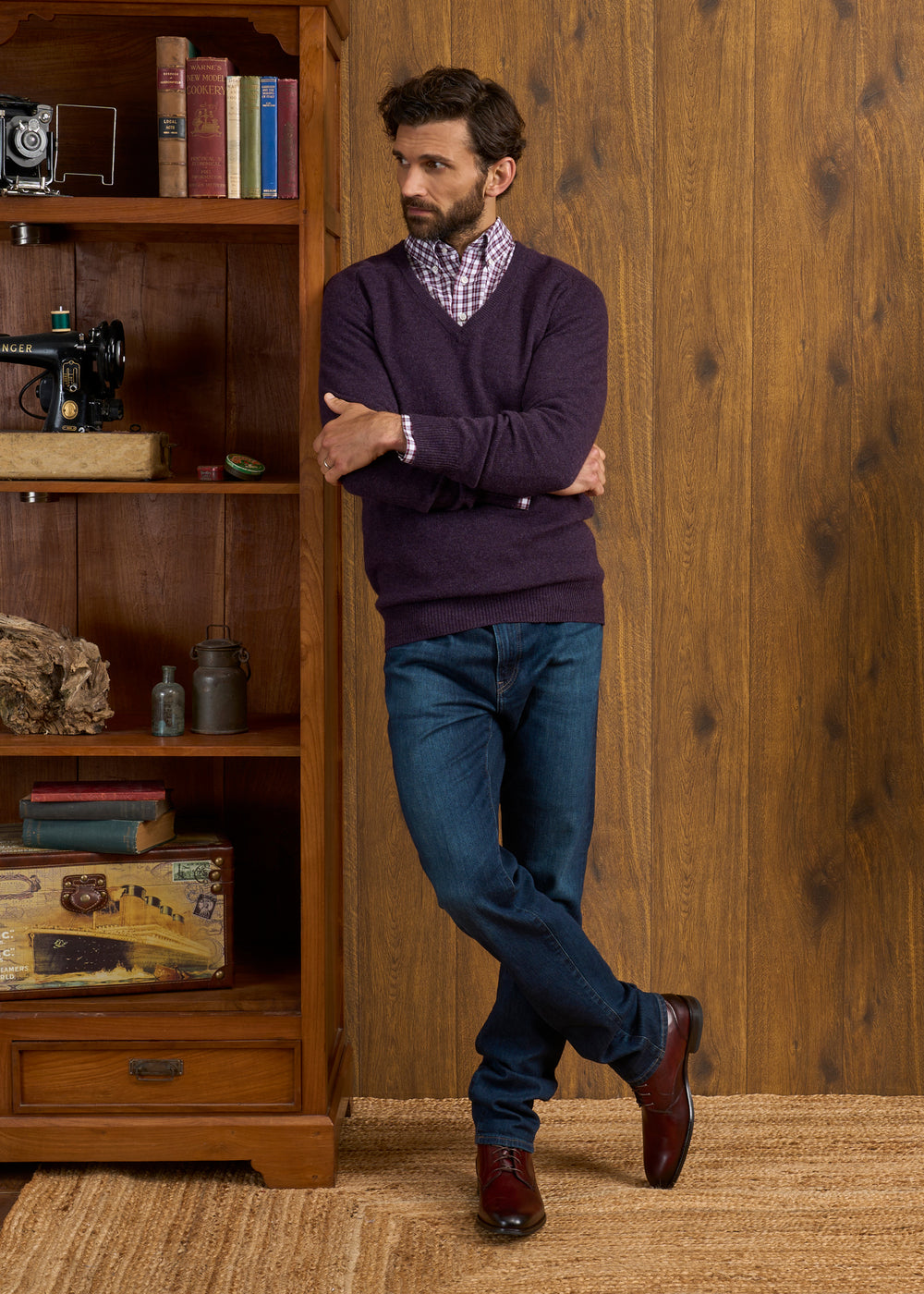 Man in Hampshire Lambswool Vee Neck Jumper (Elderberry) & jeans stands by a shelf with books, decor & vintage items.