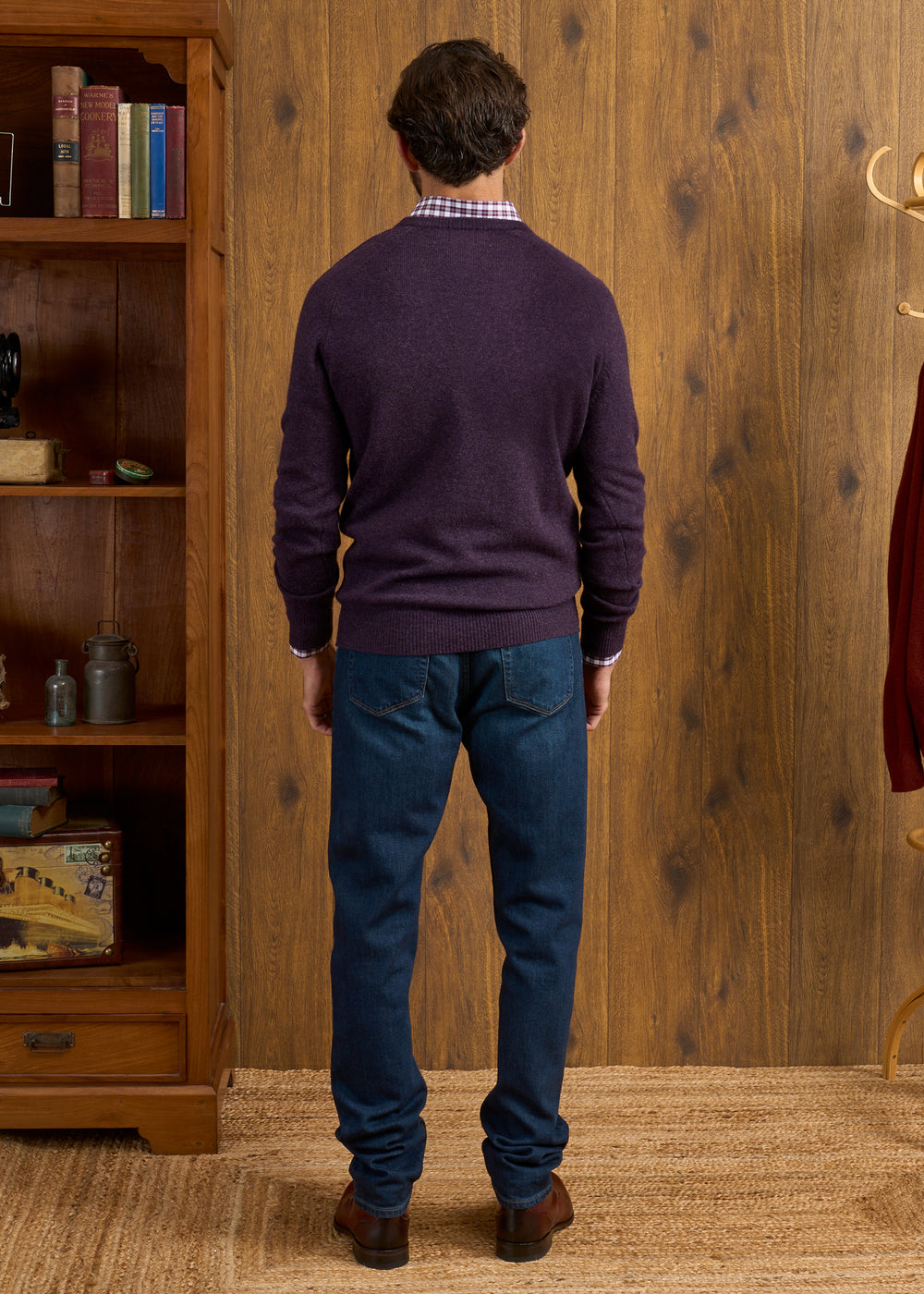 Man wears Hampshire Lambswool Vee Neck Jumper in Elderberry and jeans, facing wood wall with bookshelf and coat rack.