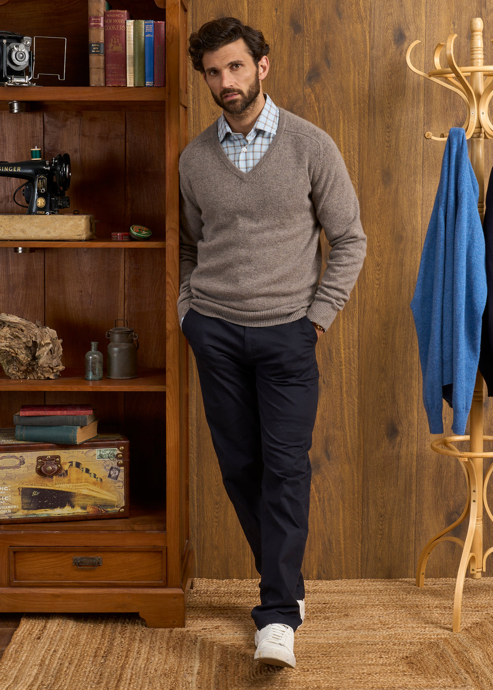 Man in Hampshire Lambswool Vee Neck Jumper (Vole), plaid shirt, navy pants, and white shoes stands by a bookcase.