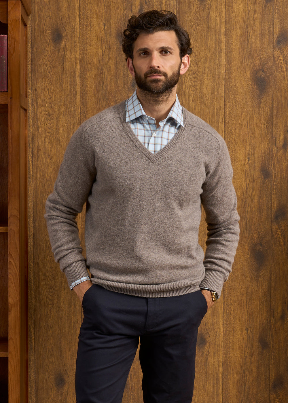 Man with curly hair wears Hampshire Lambswool Vee Neck Jumper in Vole, standing by a wooden wall with hands in pockets.