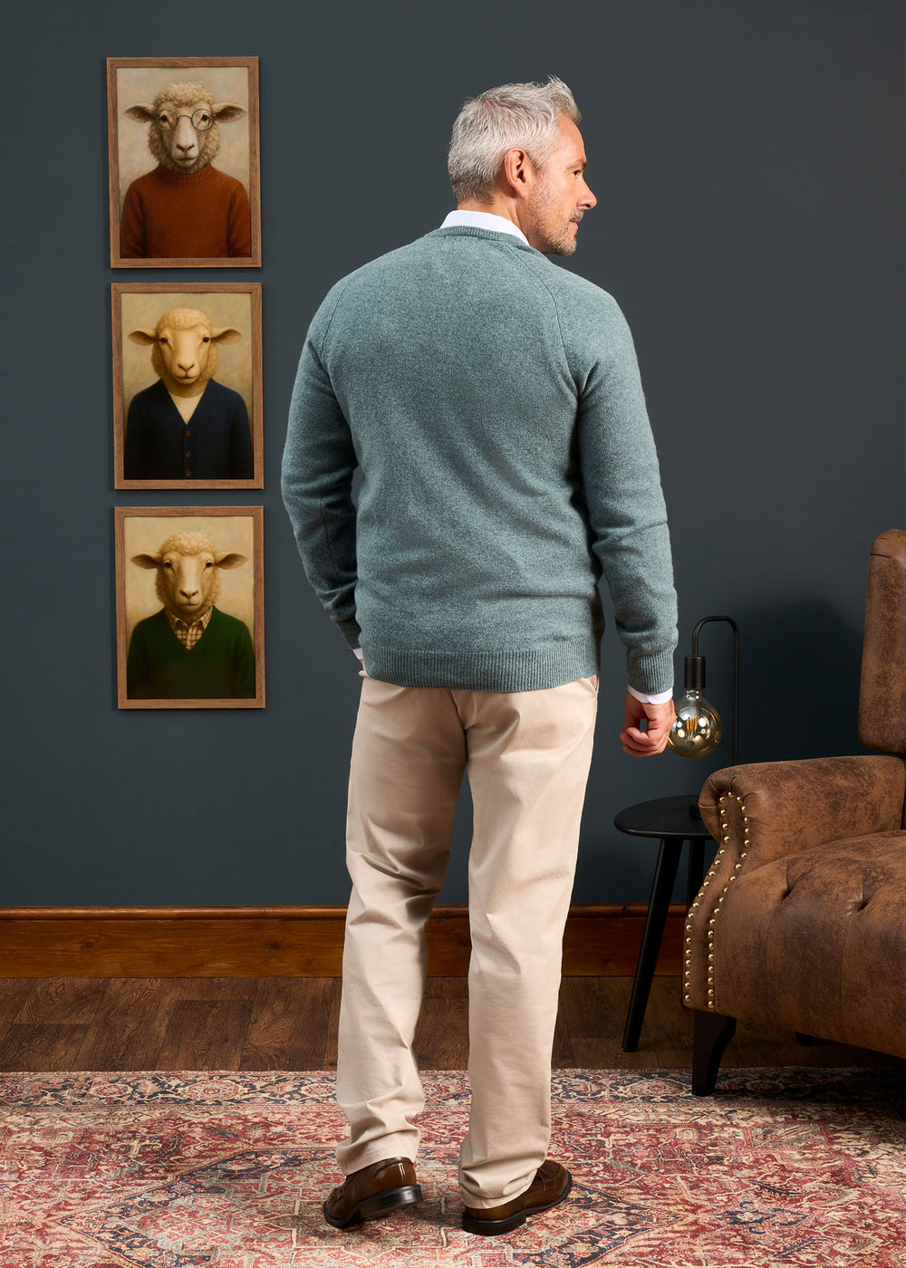 Man with gray hair in Hampshire Lambswool Vee Neck Jumper stands in a room with sheep portraits, facing away.