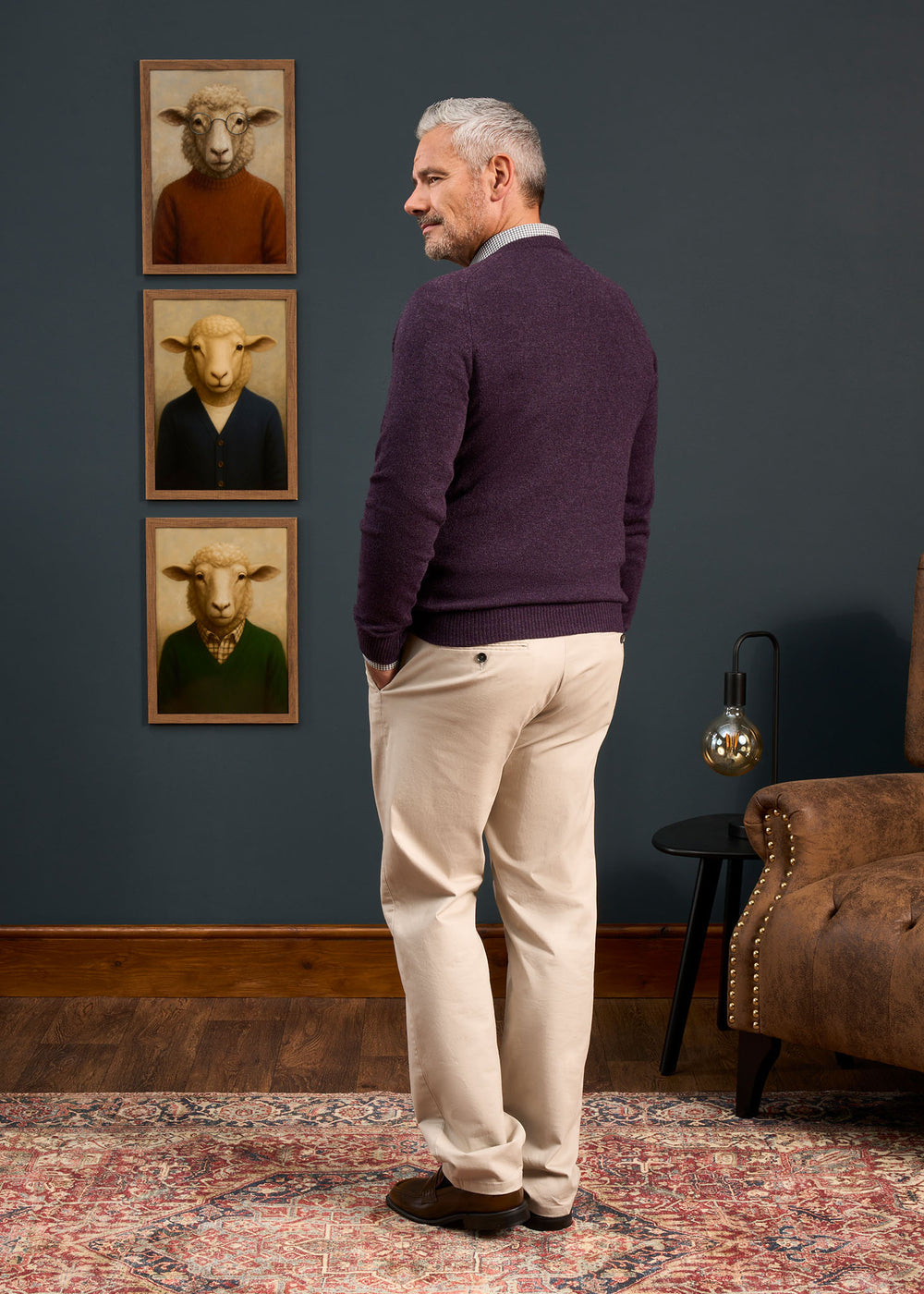 A man wears the Kilsyth Lambswool Vee Neck Jumper in Elderberry, standing sideways in a living room with sheep portraits.