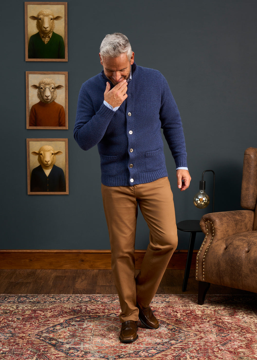 A man in a blue Landford Lambswool Buttoned Mock Neck Jumper stands indoors, sheep portraits on the wall behind him.