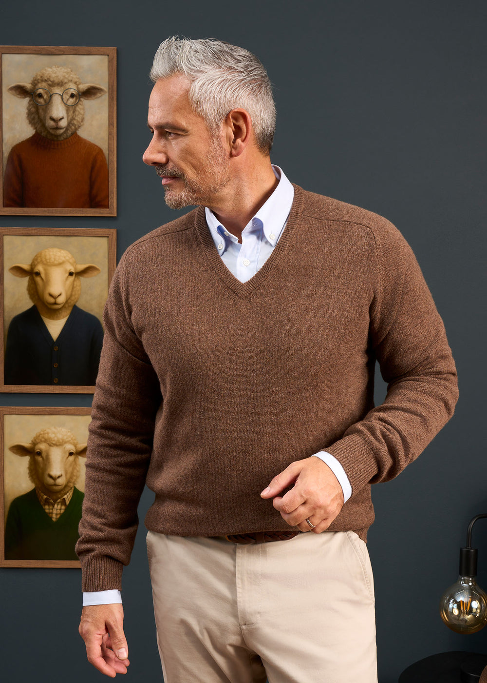 A man in the Hampshire Lambswool Vee Neck Jumper in Tobacco stands by sheep portraits, looking left against a dark wall.