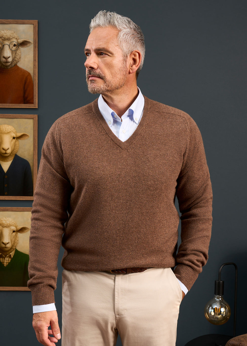 Man with gray hair wears the Hampshire Lambswool Vee Neck Jumper In Tobacco; sheep portraits and a lamp are behind him.