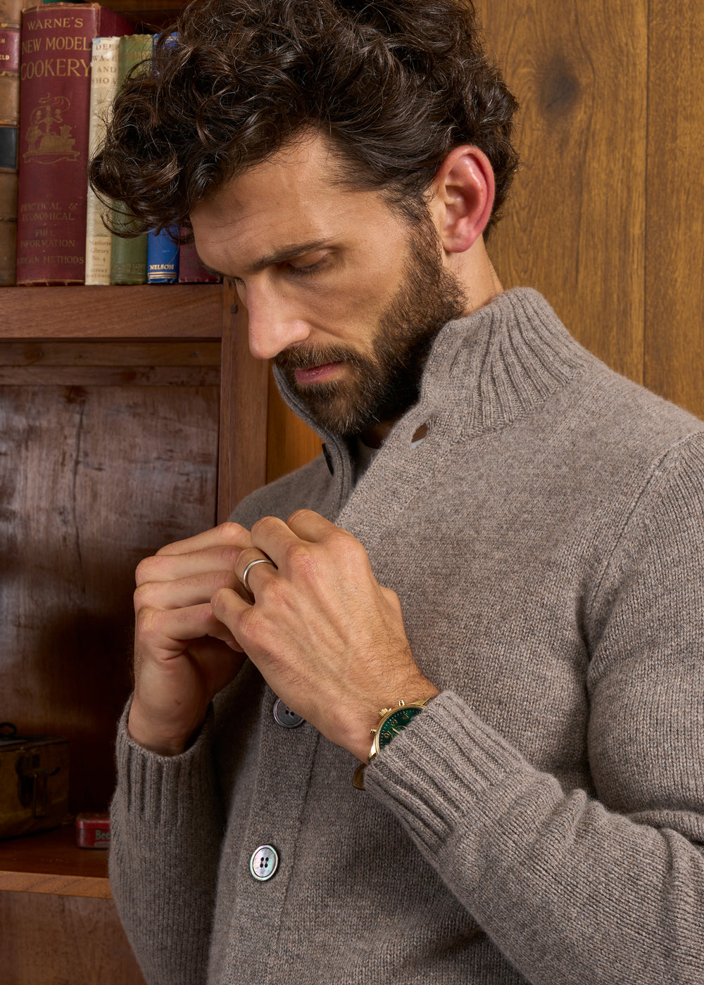 Man in Landford Lambswool Buttoned Mock Neck Jumper, gold watch, stands by bookshelf with books.