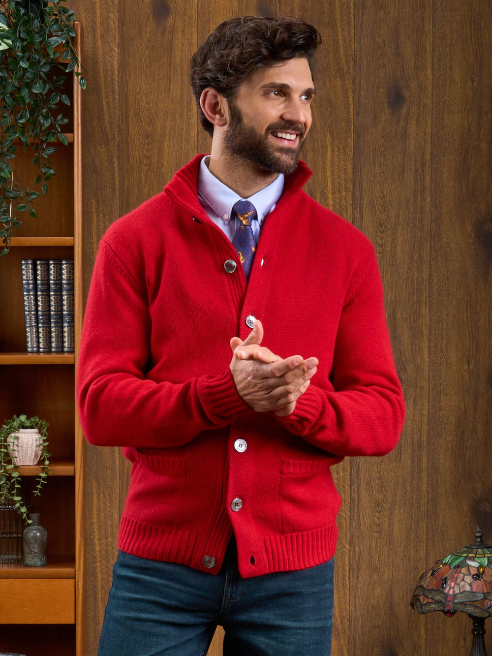 Smiling man in Landford Lambswool Mock Neck Jumper, Dubonnet, stands clasping hands; shelves and plants behind him.