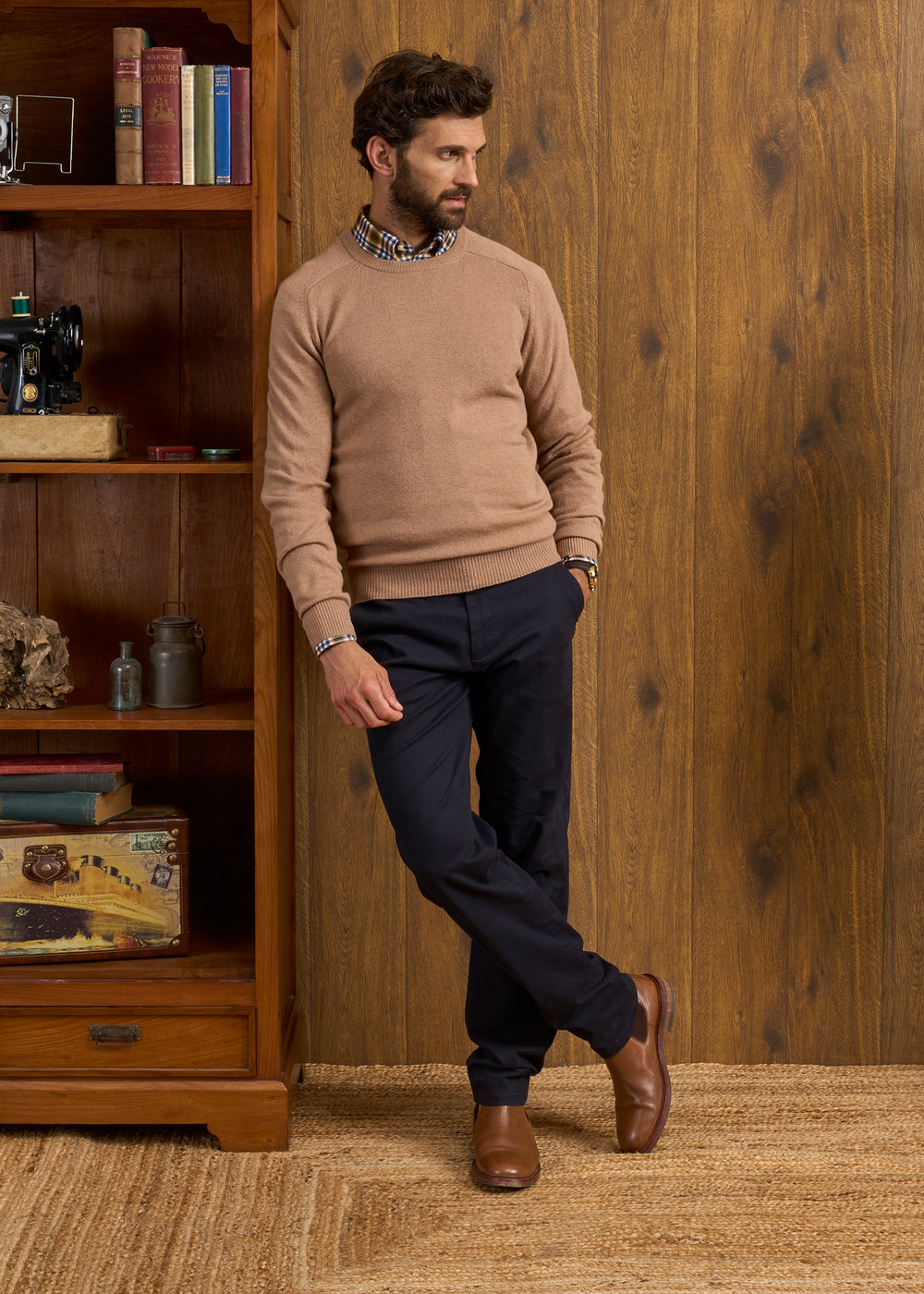 Man in Lenzie Lambswool Crew Neck Jumper in Camel, navy pants, brown shoes stands by bookcase and wood-paneled wall.
