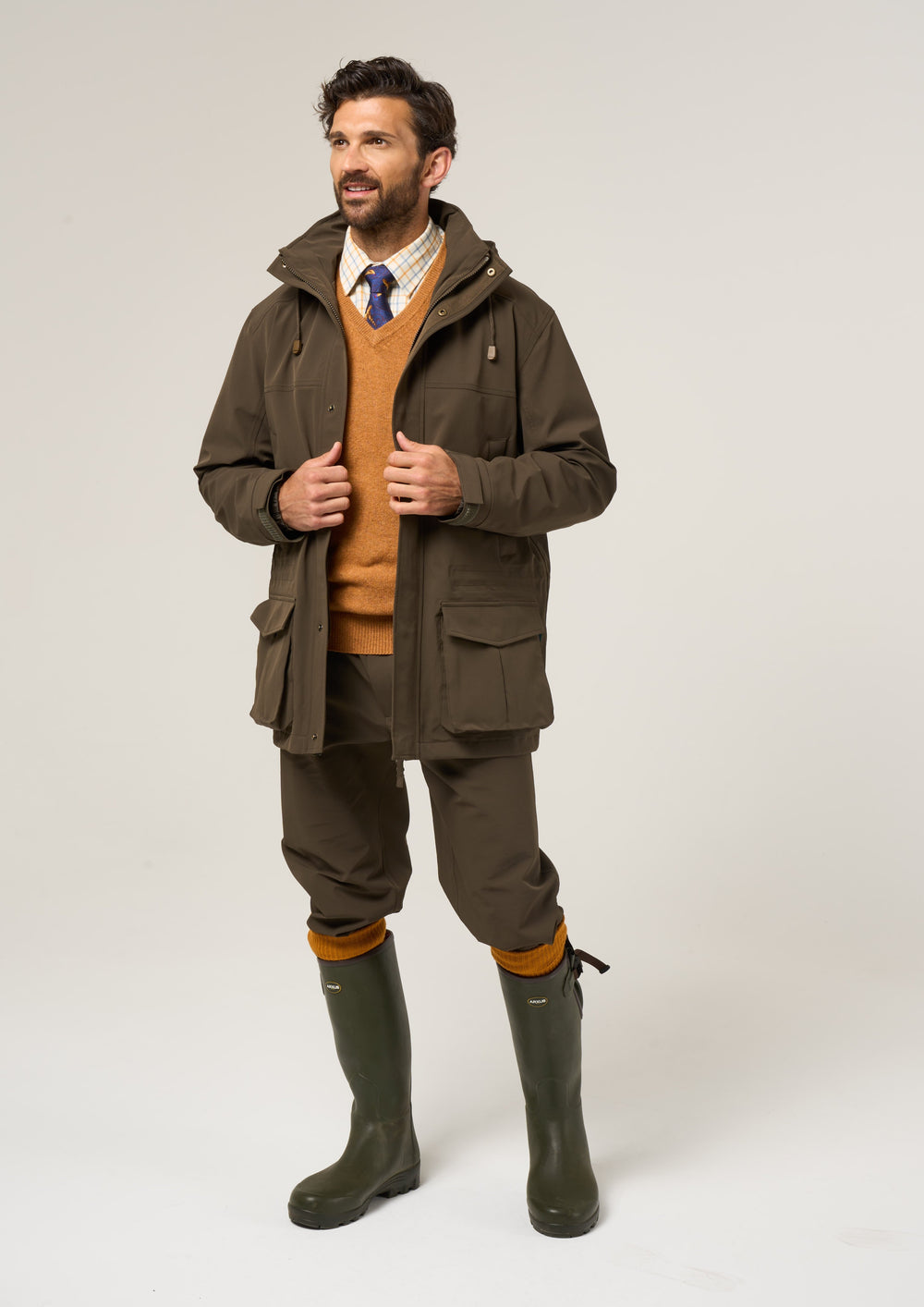 Man in a Lockwood Men’s Shooting Coat in Olive, tan sweater, dress shirt, tie, olive pants, green boots, smiling.