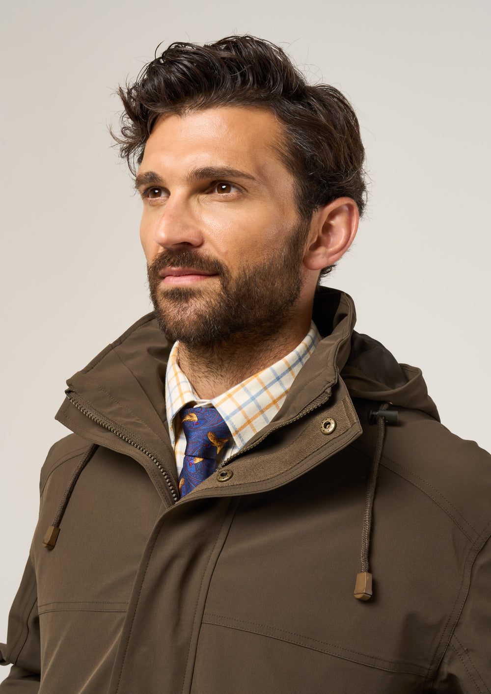 A man with brown hair and beard in a Lockwood Men’s Shooting Coat in Olive, plaid shirt, and blue tie looks aside.