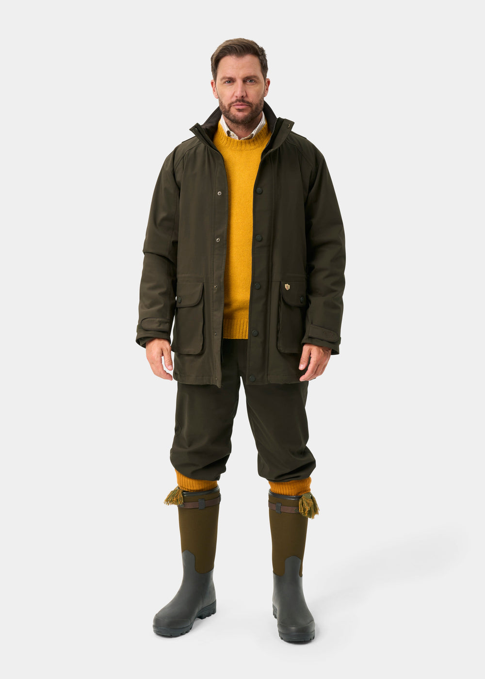 Man in Dunswell Men’s Waterproof Field Coat in Olive, yellow sweater, tassel boots, stands against white background.
