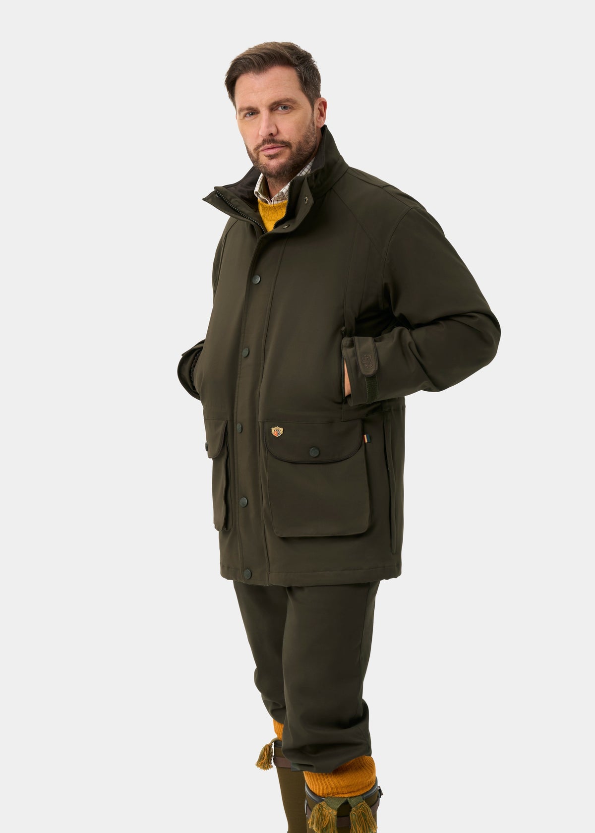 Man in Dunswell Men’s Waterproof Field Coat in olive, yellow sweater, and olive pants stands on white background.