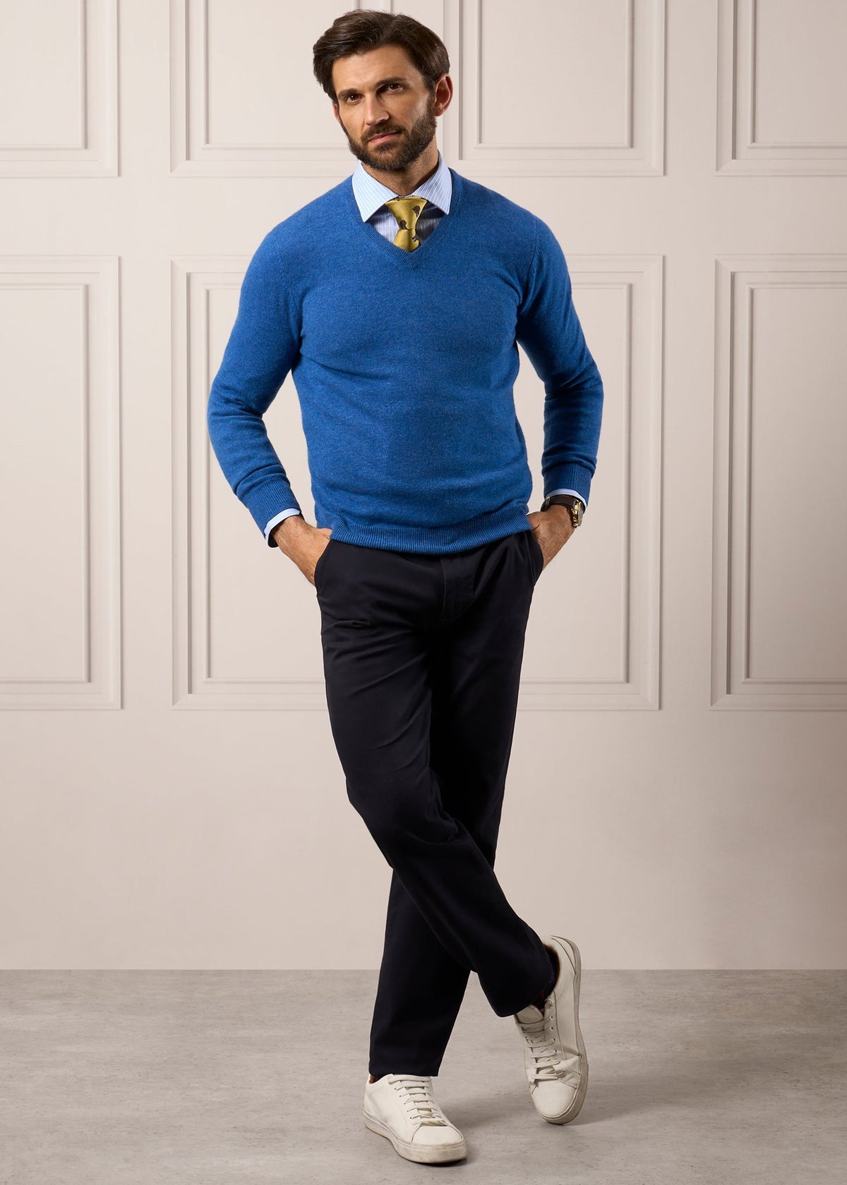 Man in Haddington Pure Cashmere V-Neck Jumper in Denim, white shirt, tie, pants, and sneakers stands by paneled wall.