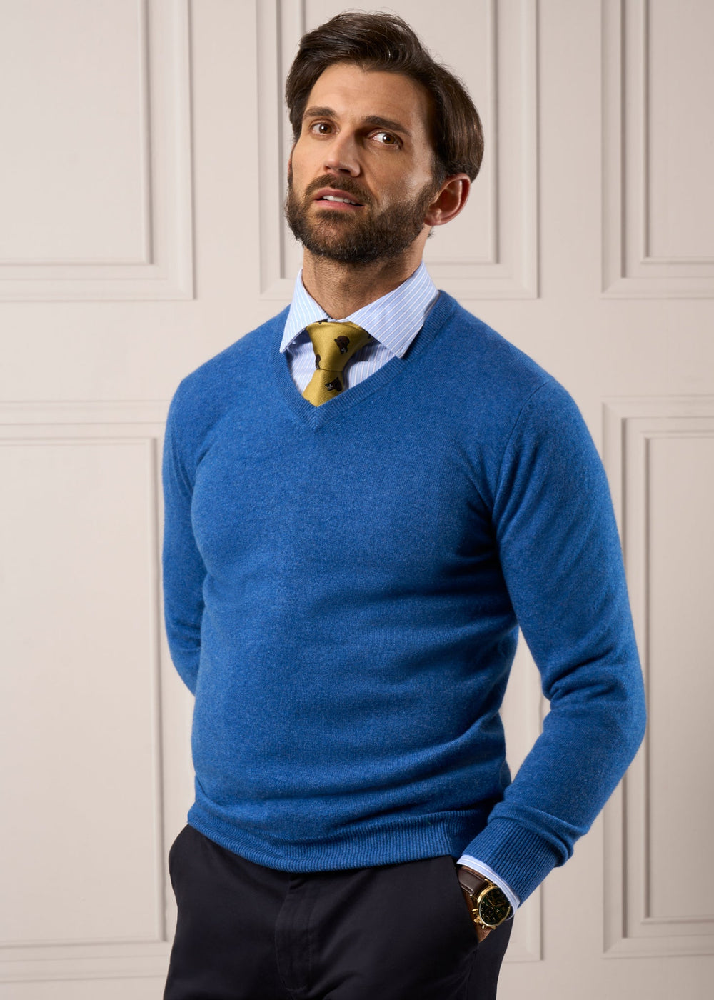 Man in Haddington Pure Cashmere V-Neck Jumper in Denim, striped shirt, and yellow tie stands by a paneled wall.