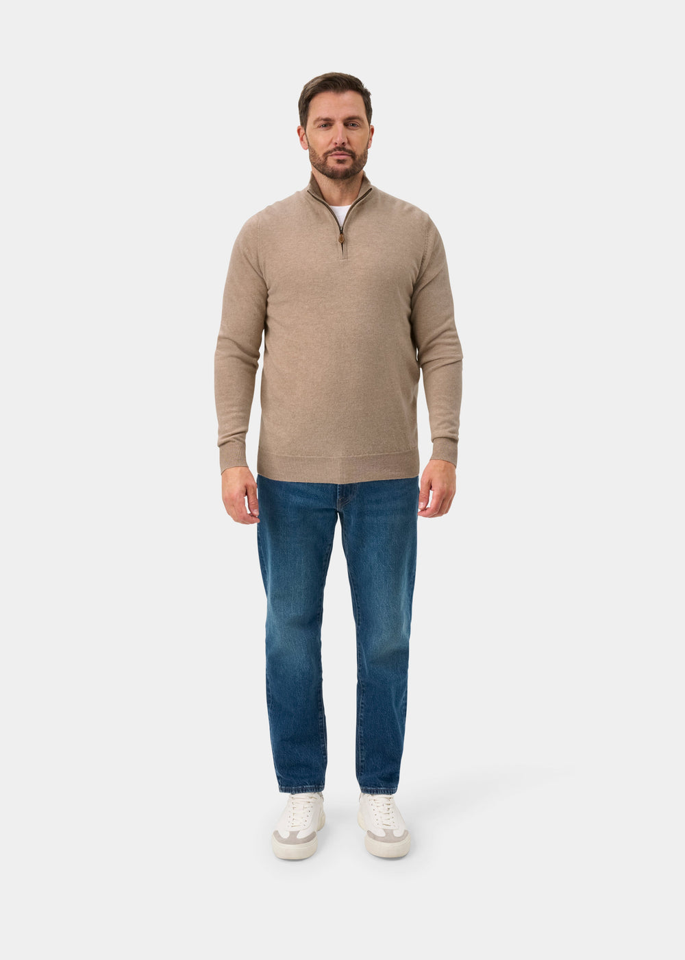 Man wears Hanbury Pure Cashmere Quarter Zip Jumper in Linen, blue jeans, and white sneakers against a plain backdrop.