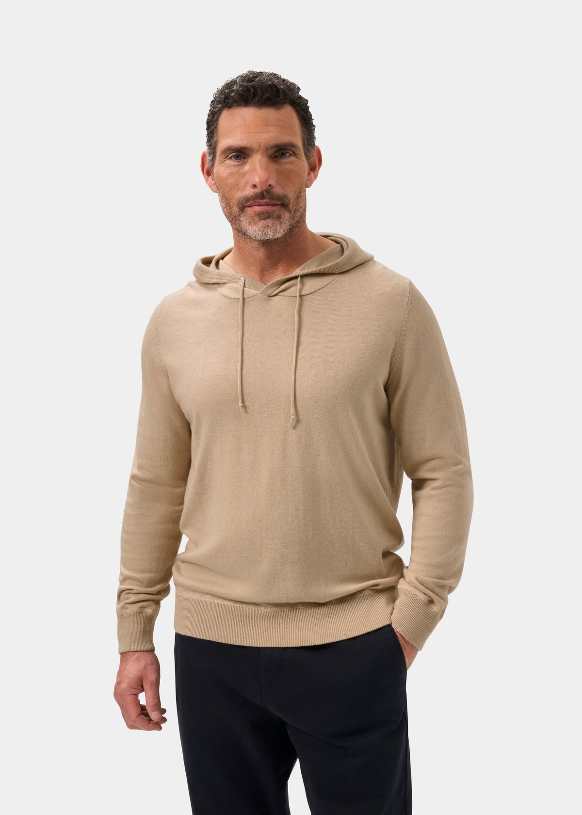 Man in Gaisby Cotton Merino Hoodie in Oatmeal, dark pants, one hand in pocket, standing against light background.