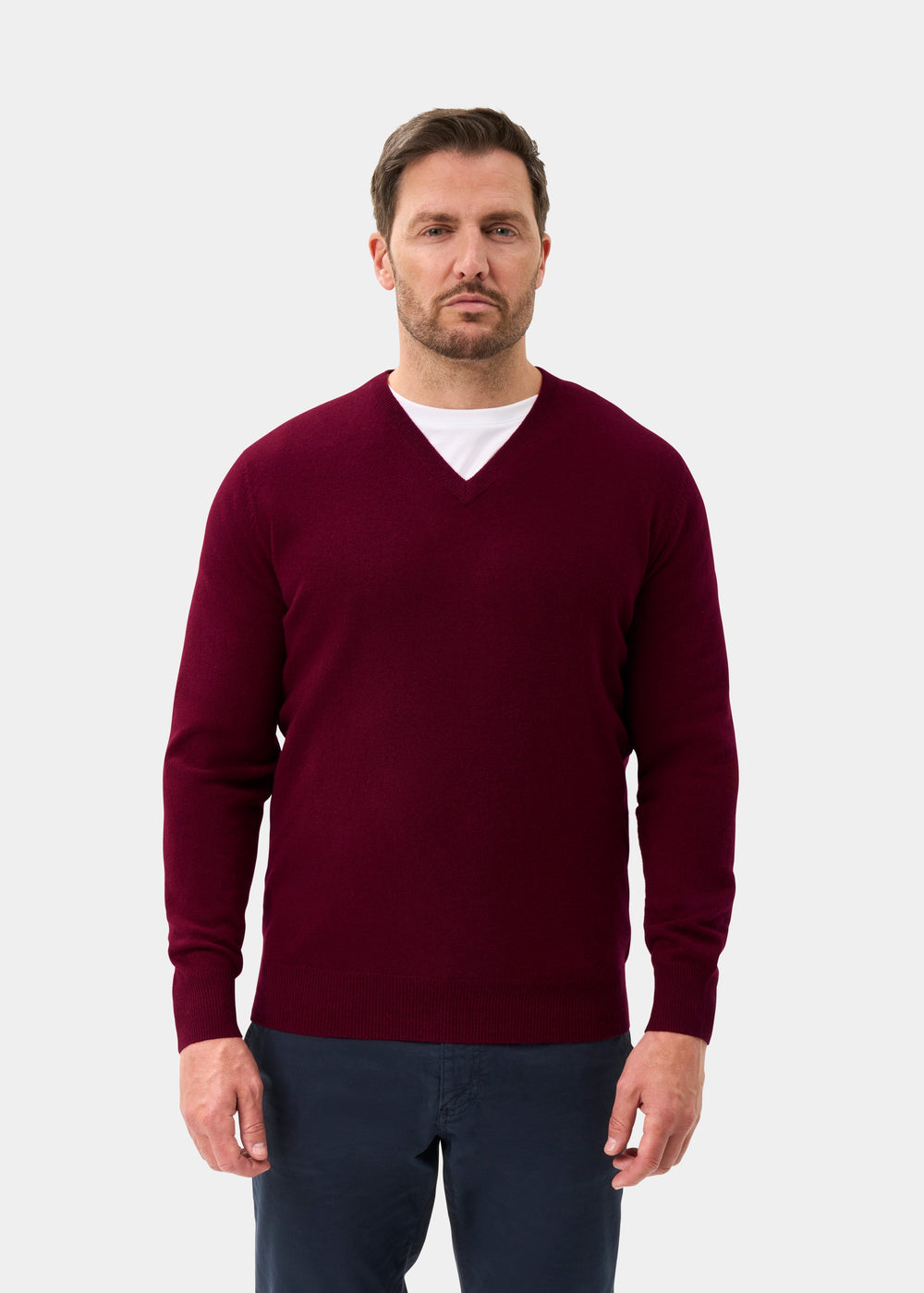 Man wears Albury Geelong Wool V-Neck Jumper in Claret over a white shirt and navy pants, on a plain light background.