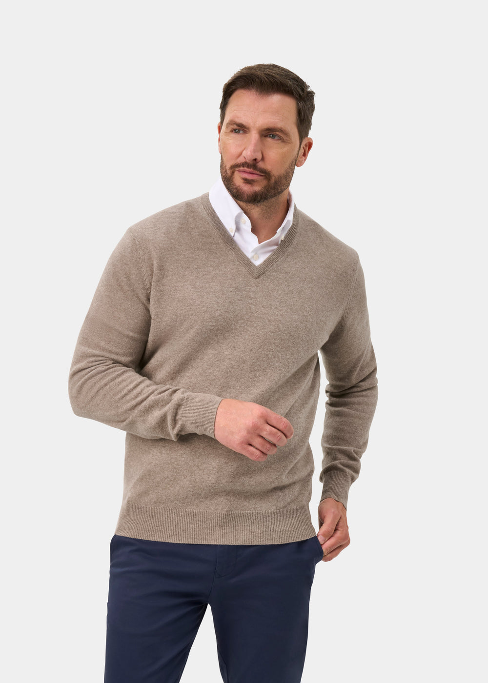 Man in Albury Geelong Wool V-Neck Jumper in Mushroom, white shirt, navy pants stands against a white background.