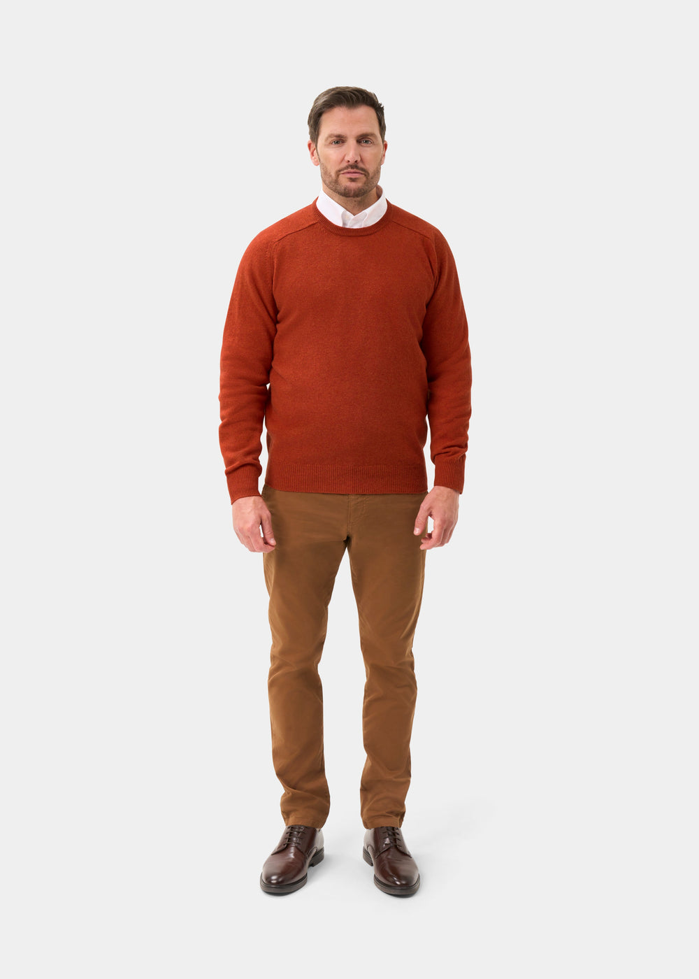 A man faces forward in a Dorset Lambswool Crew Neck Jumper in Ember, tan pants, and brown shoes against a plain background.