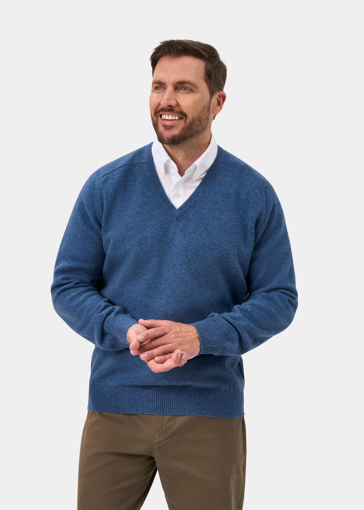 Man wears Hampshire Lambswool Vee Neck Jumper in Jeans—Classic Fit, smiling with hands clasped against plain background.