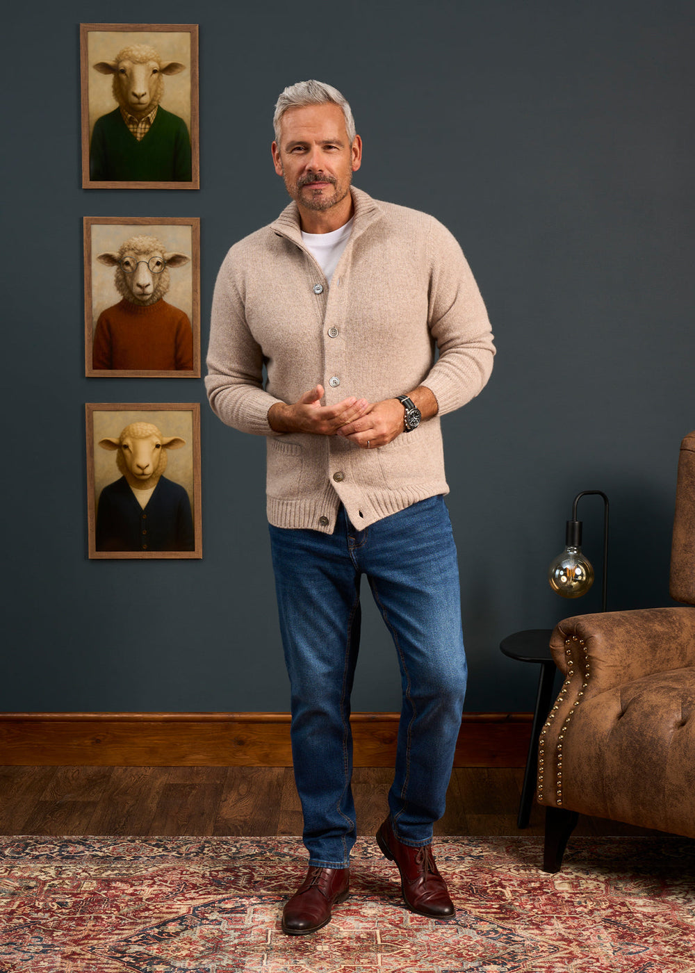 Man wears Landford Lambswool Buttoned Mock Neck Jumper in Cobble, jeans; stands by chair, sheep art behind.