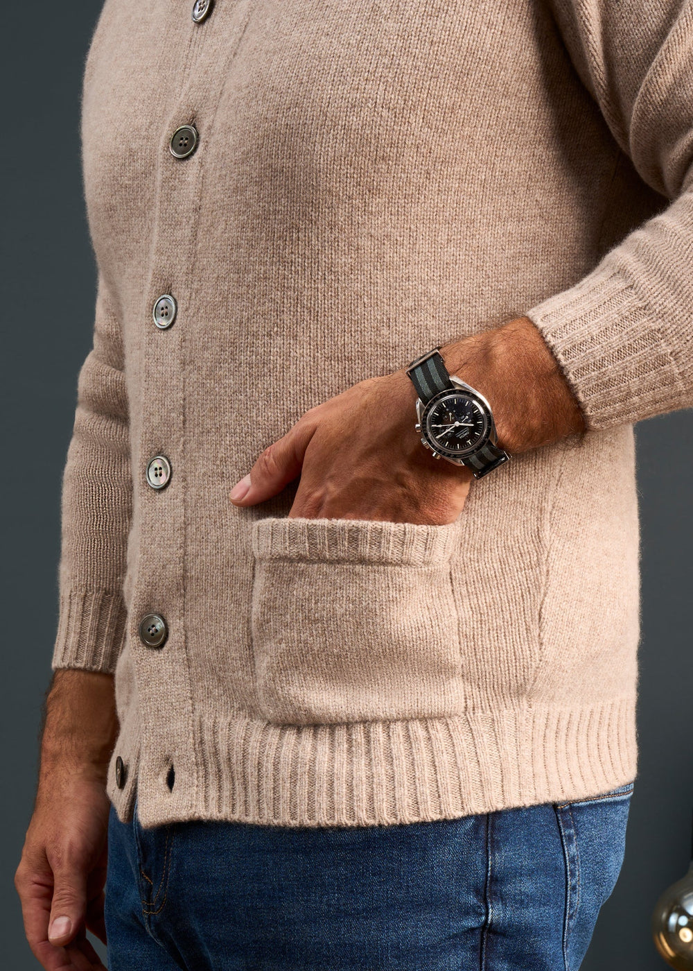 Man in Landford Lambswool Buttoned Mock Neck Jumper in Cobble and jeans, black wristwatch, dark background.