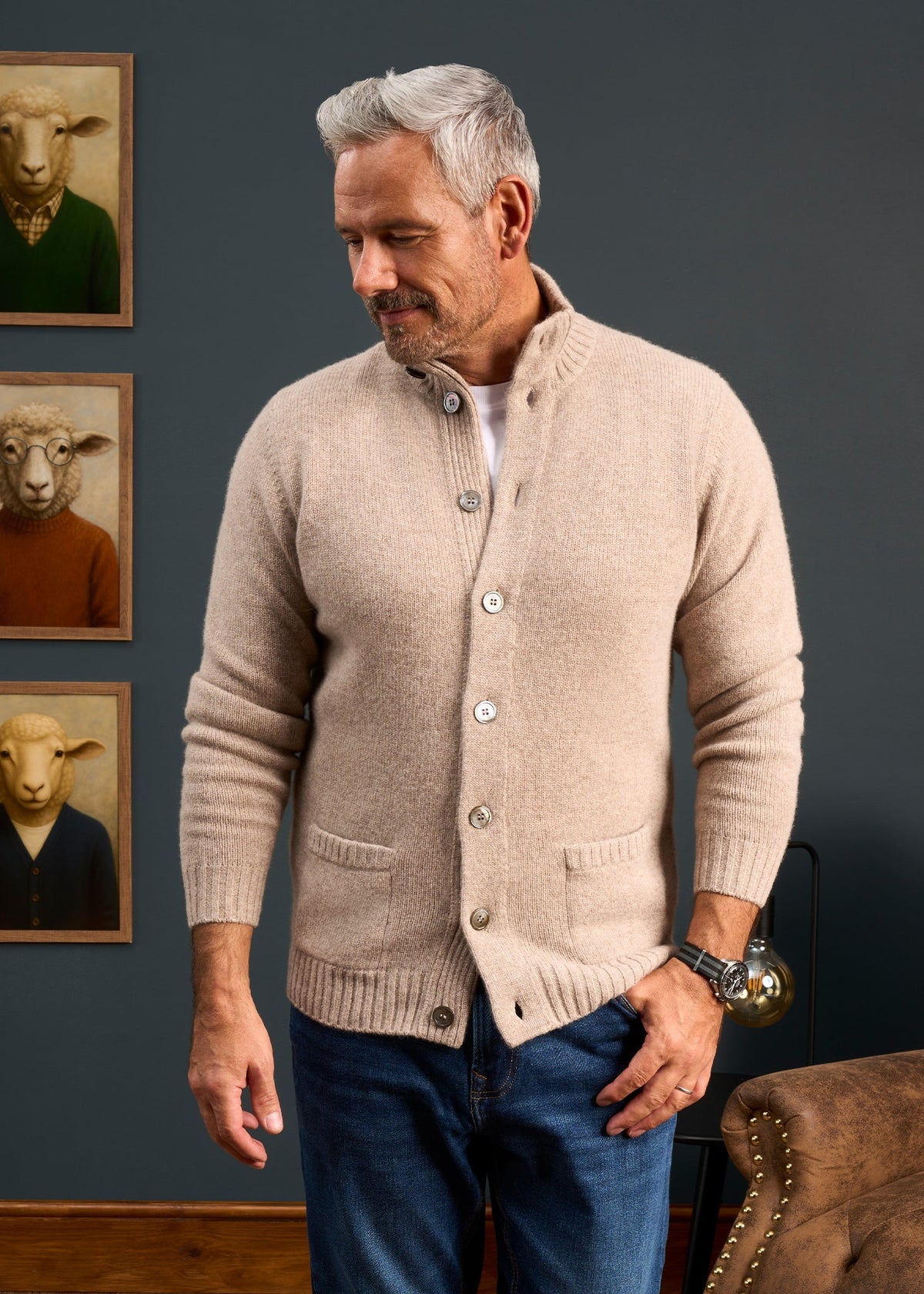 A man in the Landford Lambswool Jumper stands by a wall with three sheep portraits in sweaters, smiling down.