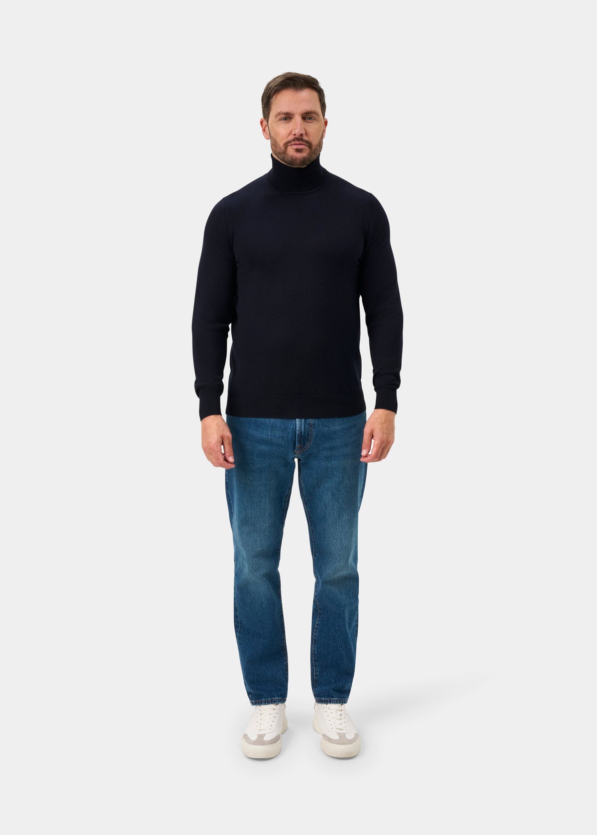 Man in a Linton Merino Wool Roll Neck Jumper in dark navy stands against a plain background, wearing blue jeans.