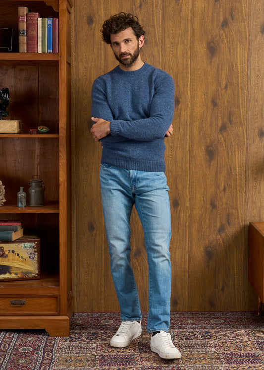 Man wears Kinnadie Supersoft Shetland Wool Denim Crew Neck Jumper, arms crossed, standing by a bookcase indoors.