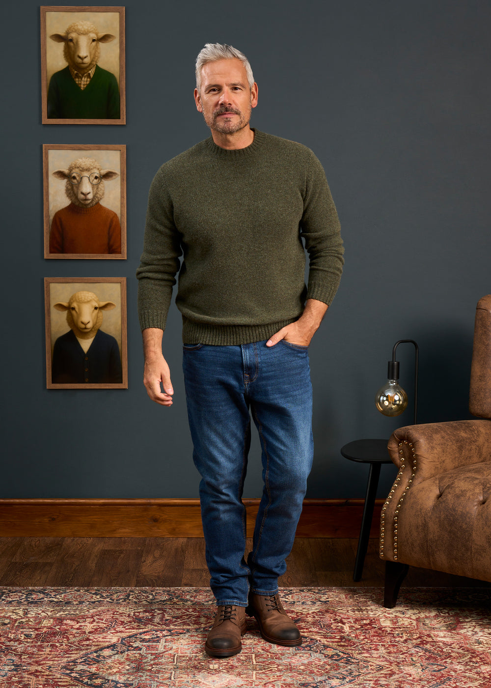 A man in a living room with sheep art wears the Kinnadie Shetland Wool Crew Neck Jumper in Moorland, jeans, and brown shoes.
