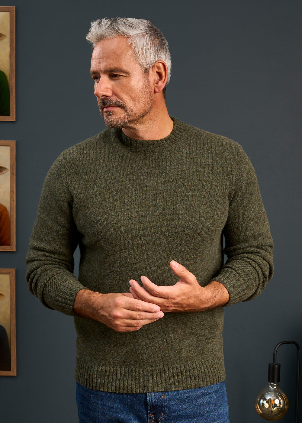 Gray-haired man in Kinnadie Supersoft Shetland Wool Crew Neck Jumper, regular fit, stands indoors by framed art.