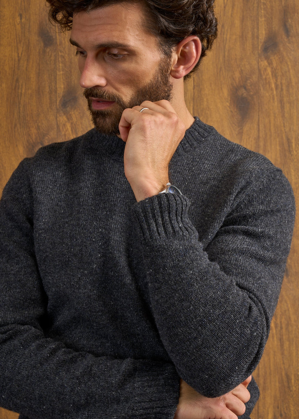 Man with curly hair wears the Kinnadie Supersoft Shetland Wool Crew Neck Jumper in Oxford Pine, standing by wood.