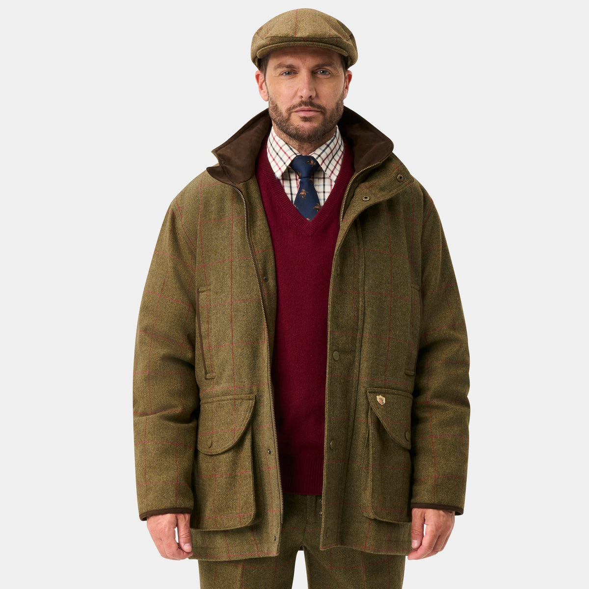 A man stands against a light gray studio background wearing an olive green hunting jacket, matching trousers, dark green rain boots, a burgundy sweater, and a brown flat cap.