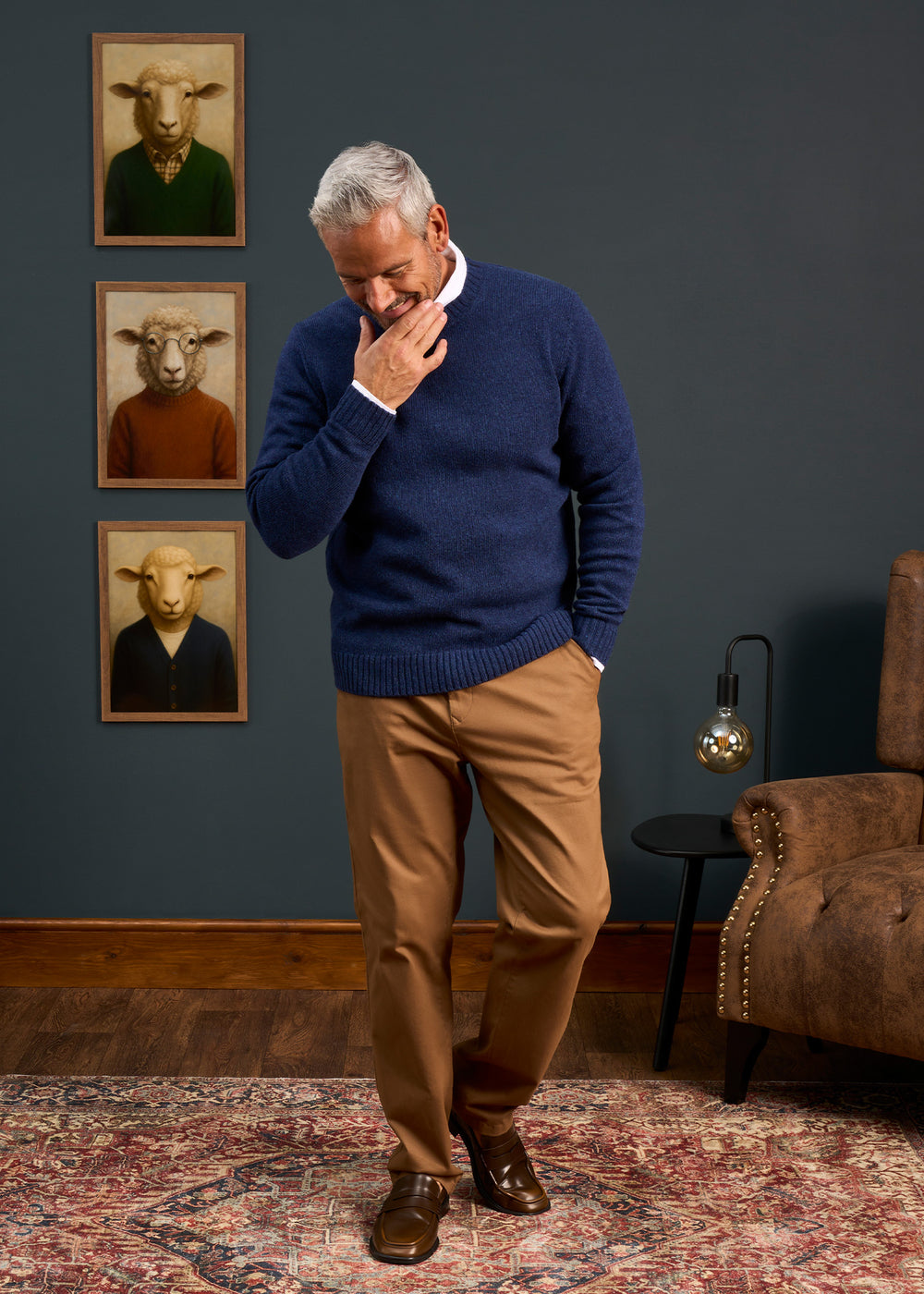 A man in a Kinnadie Supersoft Shetland Wool Crew Neck Jumper stands pensively in a cozy room with sheep portraits.