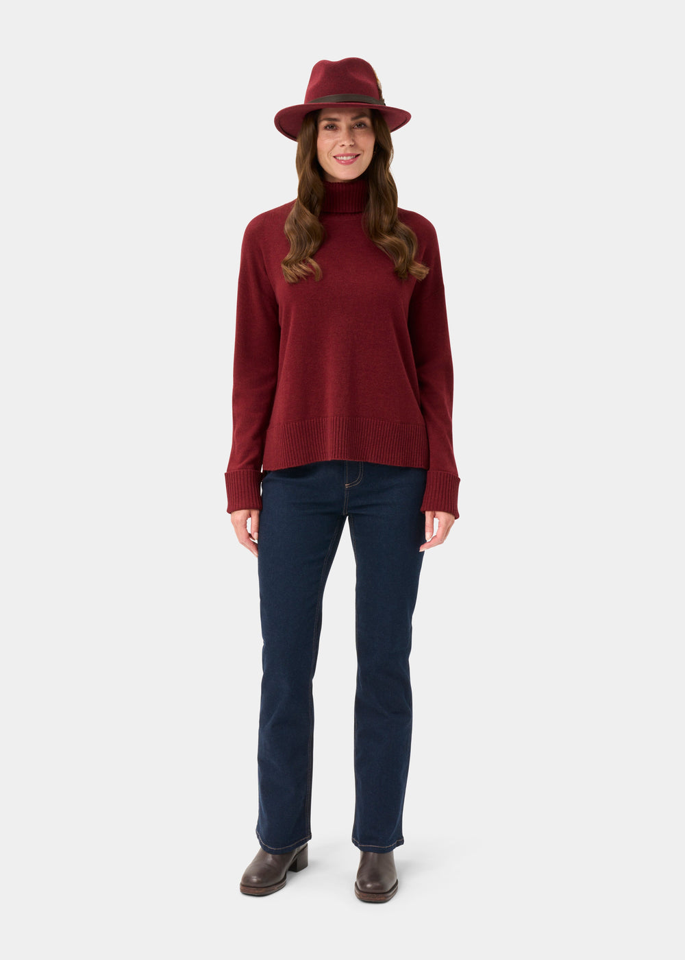 Woman in Alexa Ladies Berry Roll Neck, red hat, blue jeans, and brown boots smiles against a white background.