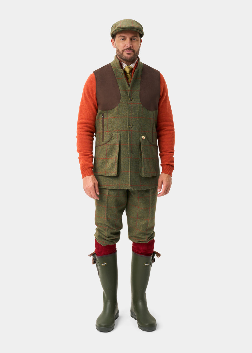 Man in Combrook Men’s Tweed Water Repellent Shooting Gilet (Reed), green tweed, brown vest, orange sweater, boots, cap.