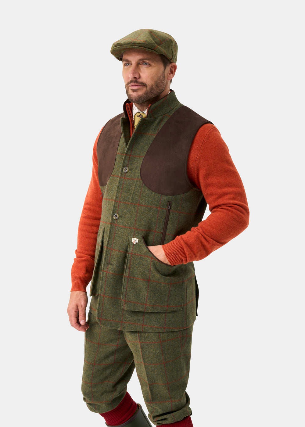 Man wears Combrook Men’s Tweed Water Repellent Shooting Gilet in Reed and orange sweater, posed on white background.
