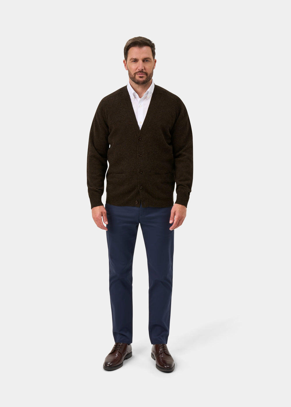 Man in Cornwall Lambswool V-Neck Cardigan in Cocoa, white shirt, blue pants, brown shoes on plain light background.