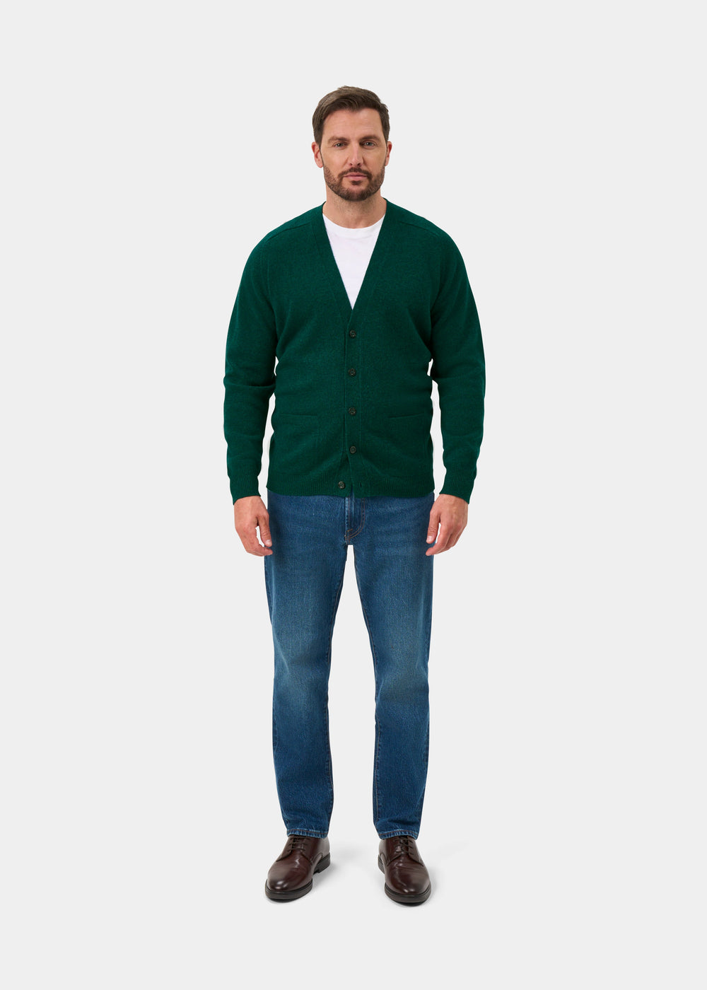 Man in Cornwall Lambswool V-Neck Cardigan in Mallard, white shirt, blue jeans, brown shoes stands against white background.